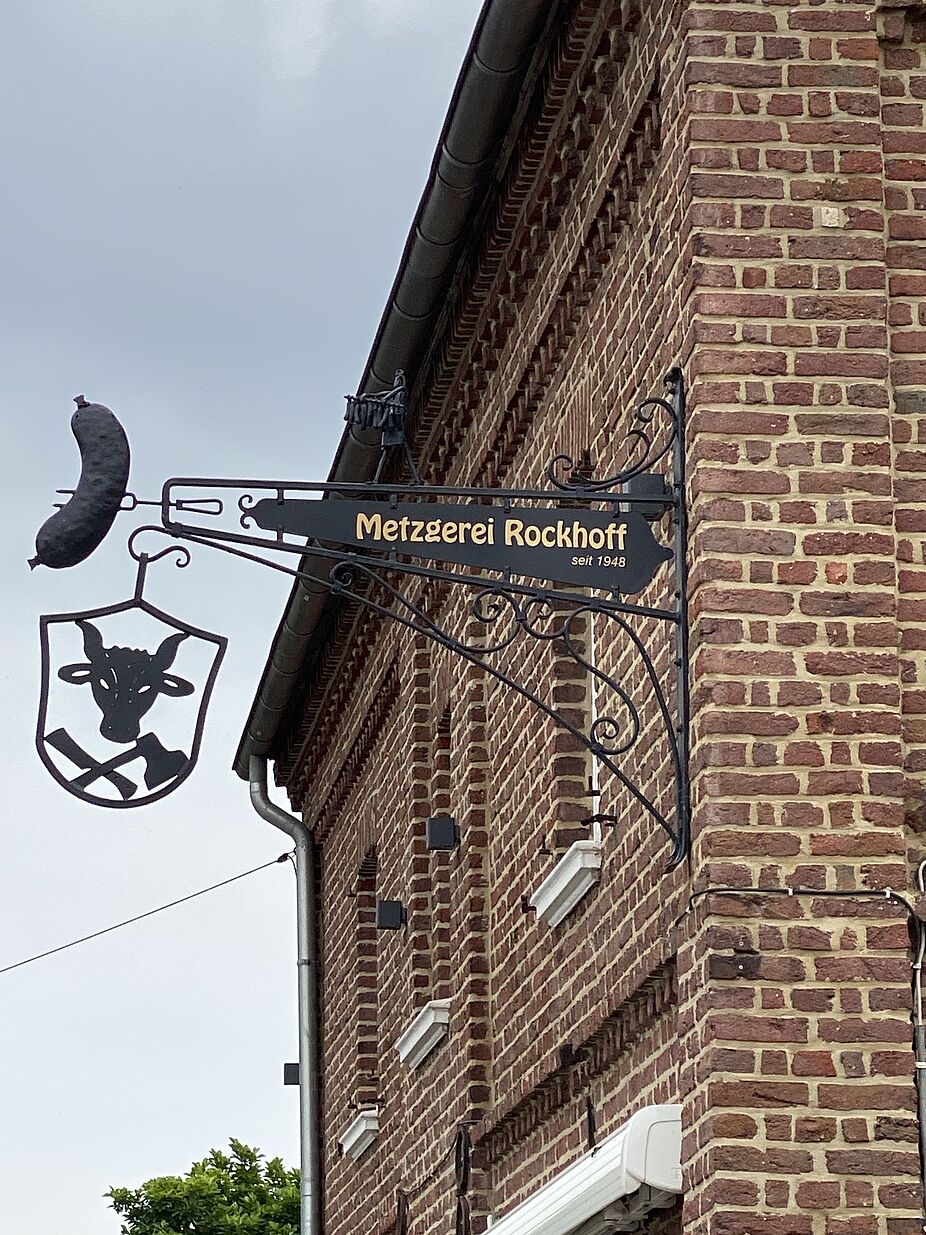 Hier wartet man gerne Hier wartet man gerne, Firmenschild der Metzgerei Rockhoff in Eppinghoven