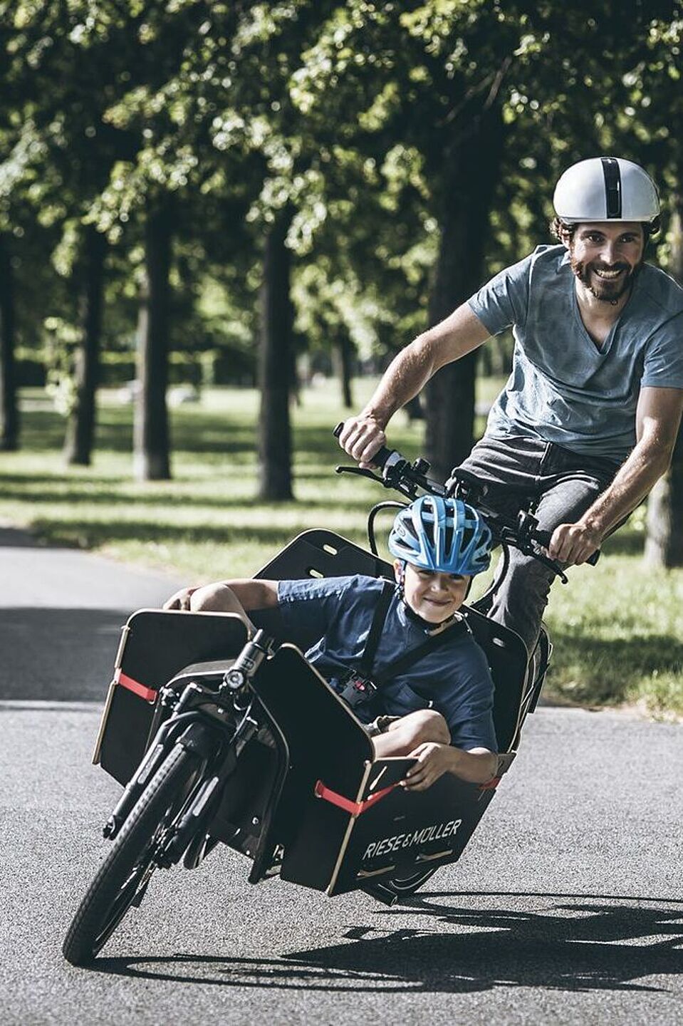 Mit Long John-Lastenrädern kann man auch sportlich fahren. Ein Mann fährt mit einem Kinder in einem Lastenrad sportlich um eine Kurve im Park